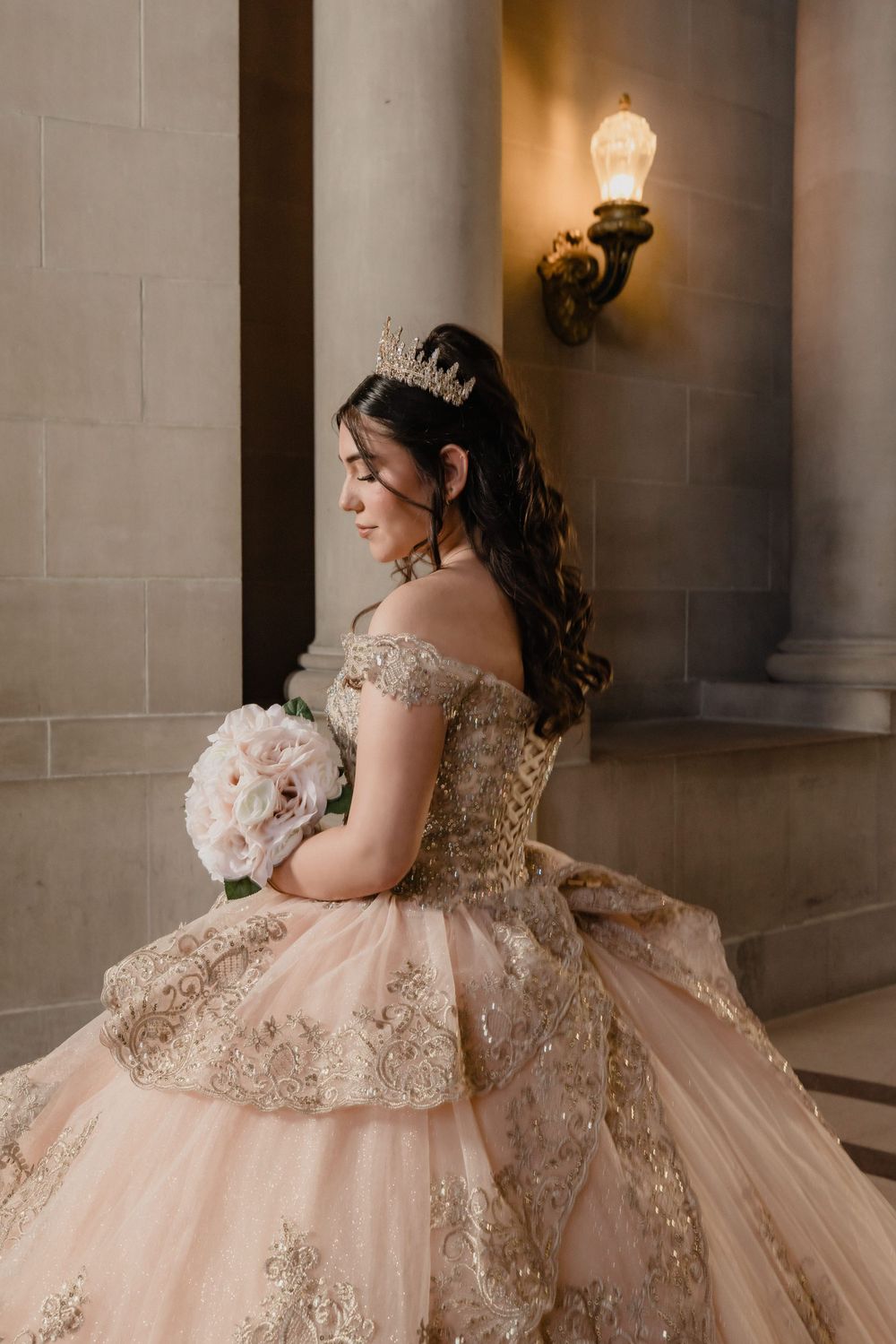 Young woman in a blush pink and gold quinceañera ball gown with tiara holds white bouquet by ornate columns.