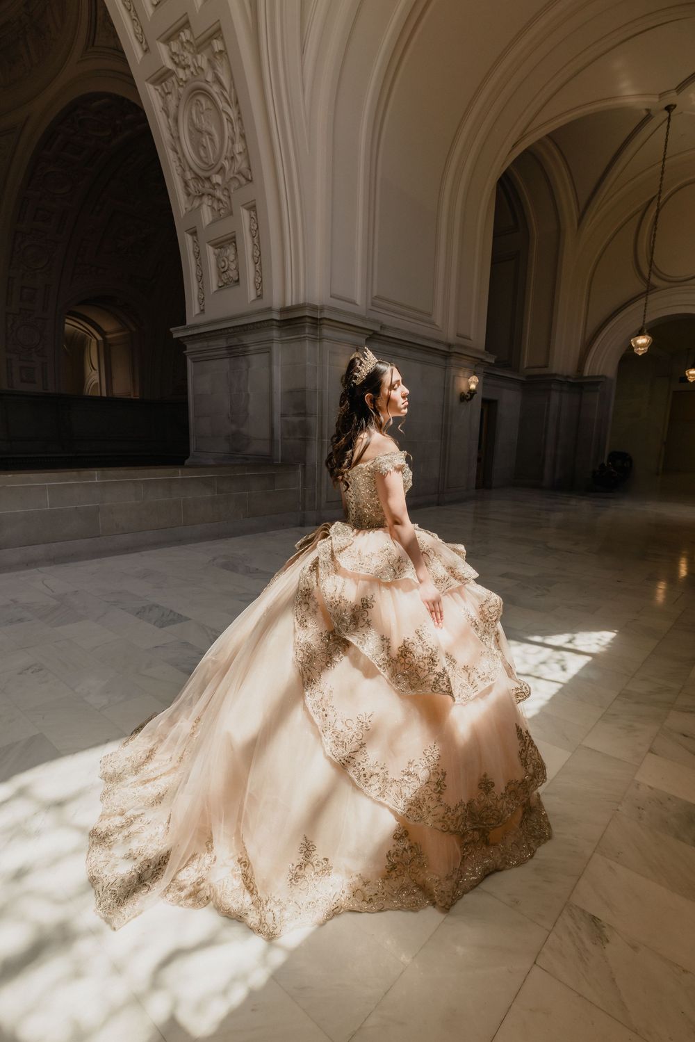 A bride in an ornate gold-embroidered ballgown poses gracefully in a sunlit marble hall.