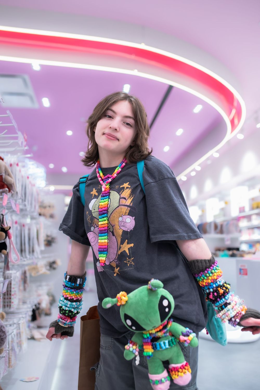 Teen in graphic tee holding green plush toy, wearing colorful kandi bracelets in a neon-lit novelty shop.
