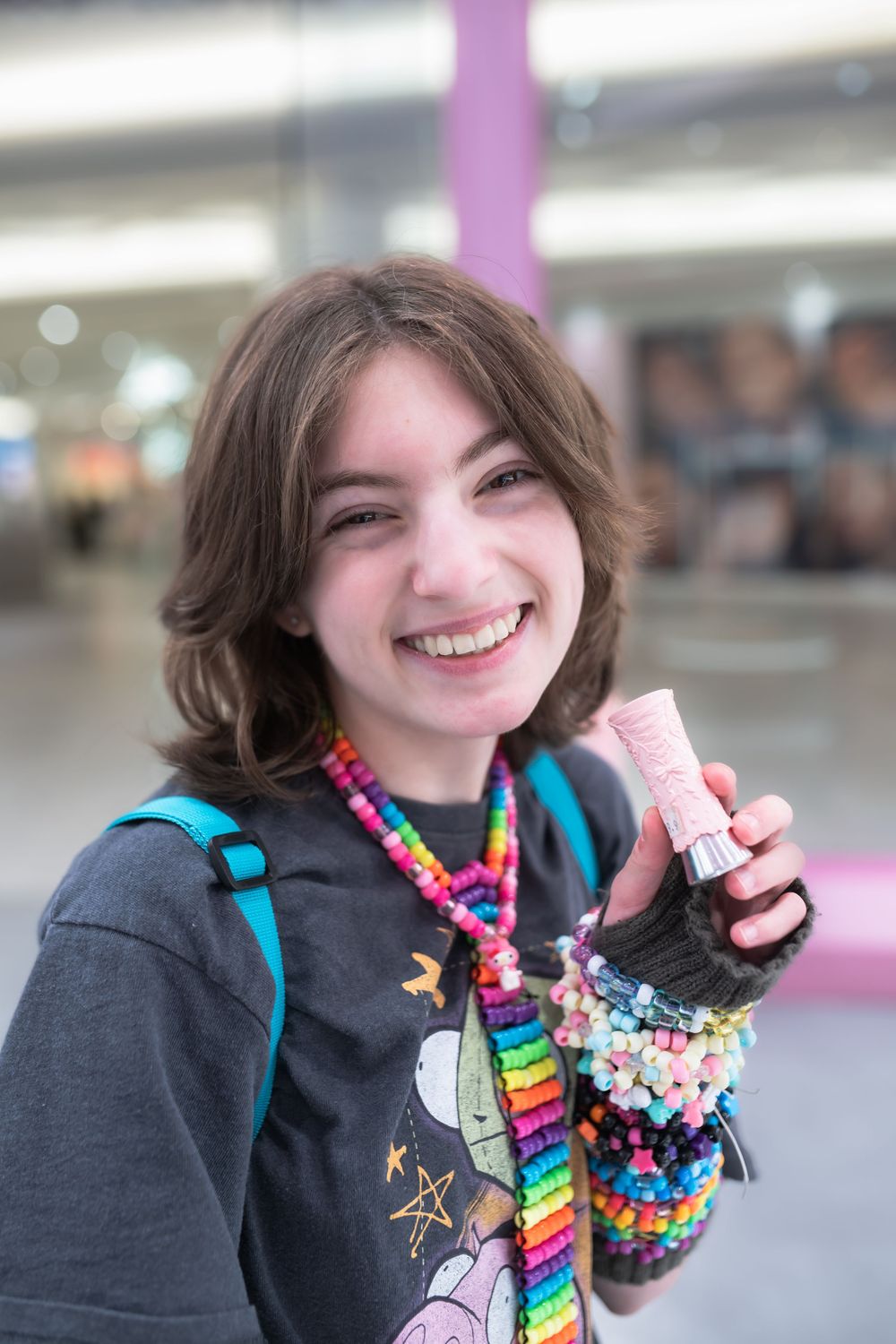 Smiling young girl wearing colorful bead necklaces and bracelets, holding a pink item, with a blue backpack.