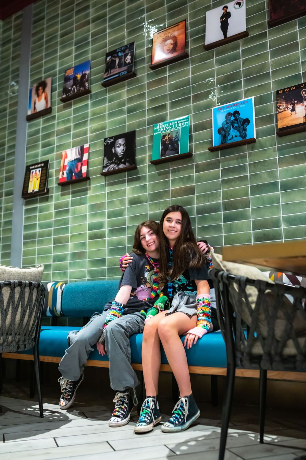 Two teens sitting on a blue bench against a green tiled wall decorated with vintage music album covers.