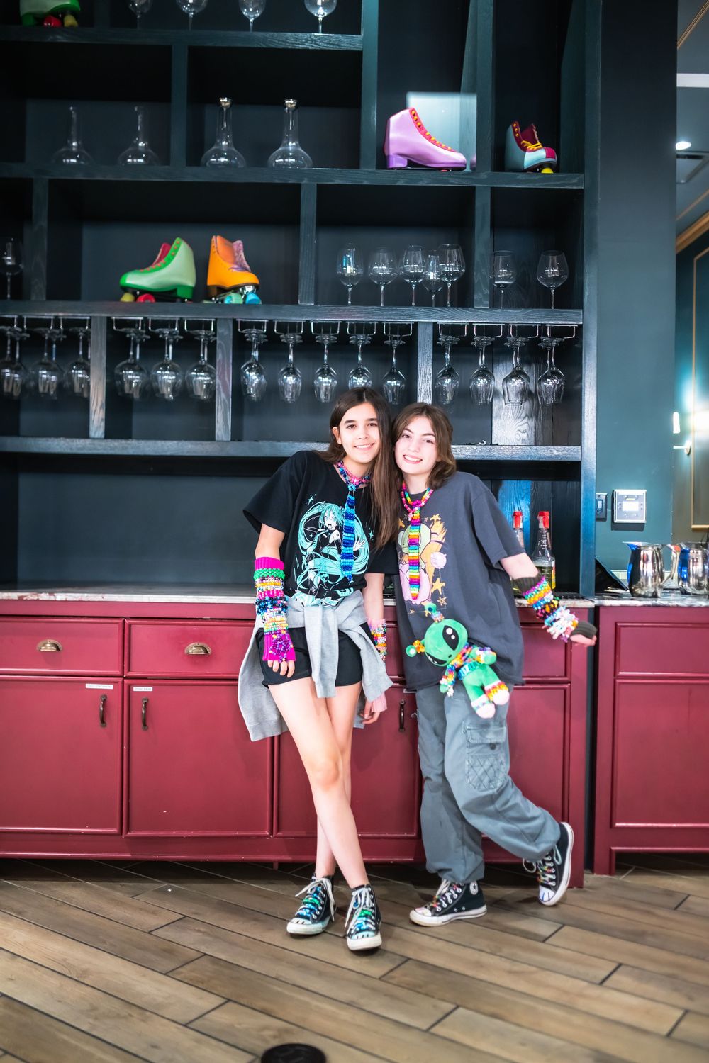 Two young women in colorful 80s-themed outfits pose in front of shelves with roller skates and wine glasses.
