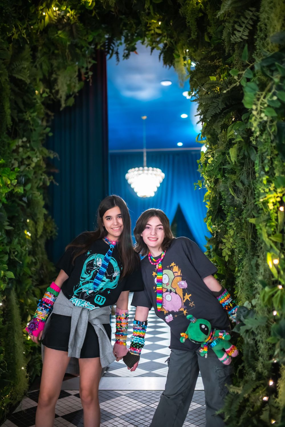 Two girls in colorful graphic tees pose under a lush green archway with fairy lights at an event.