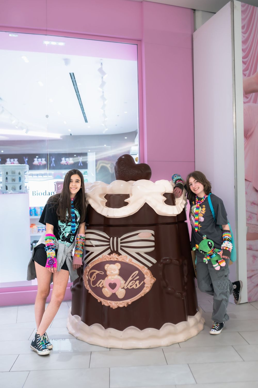 Two people posing beside a giant chocolate cake sculpture outside a Candylés store in a shopping mall.