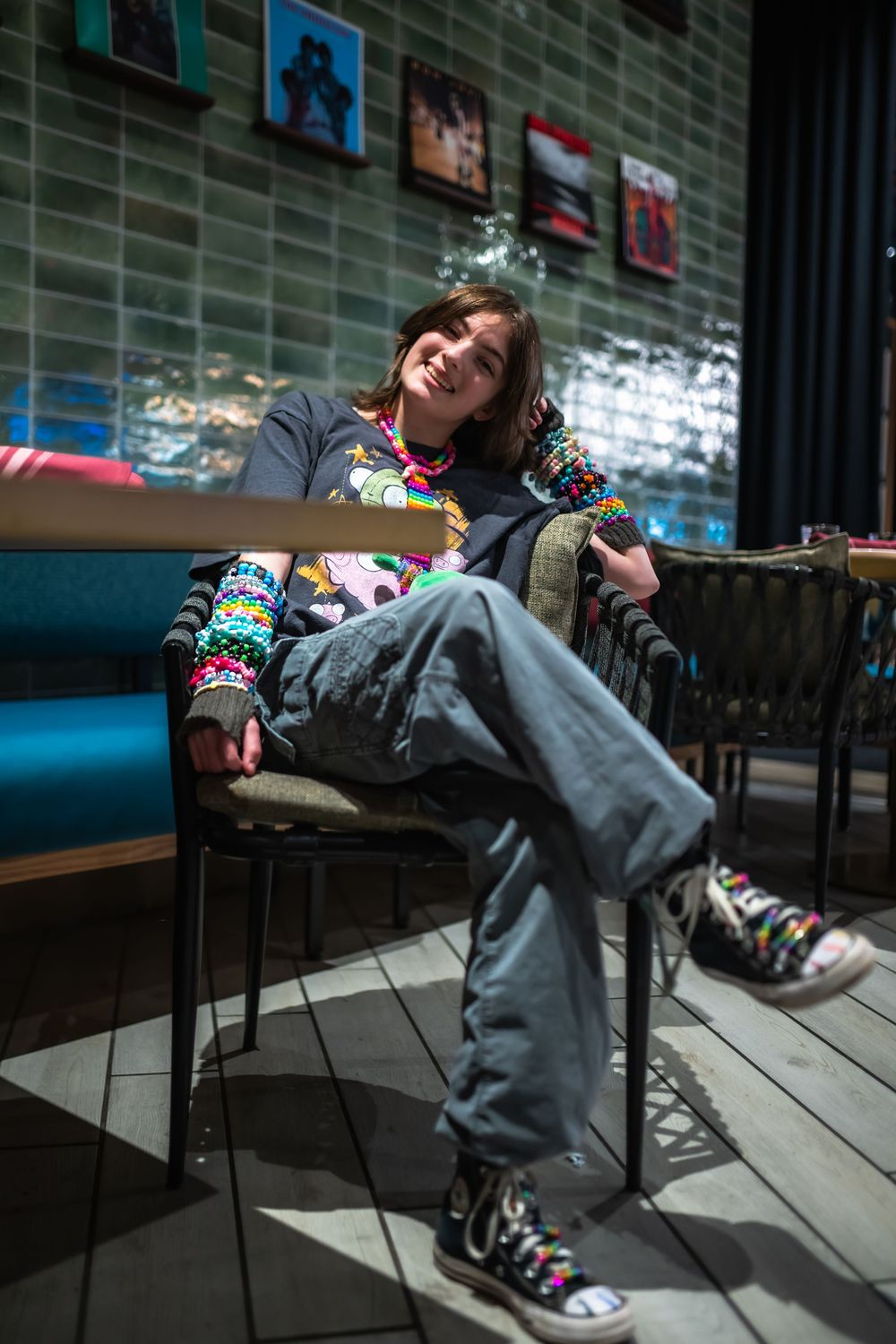 Young person sitting relaxed in chair wearing colorful bracelets, graphic tee, gray jeans, and Converse sneakers in a cafe.