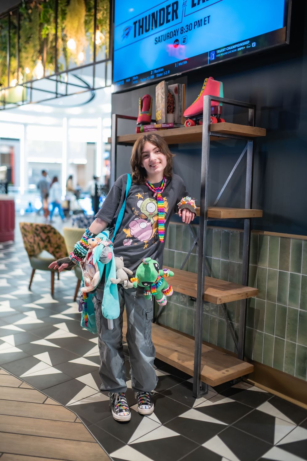 Young woman in colorful accessories and graphic tee posing by a wooden shelf display inside a modern mall.