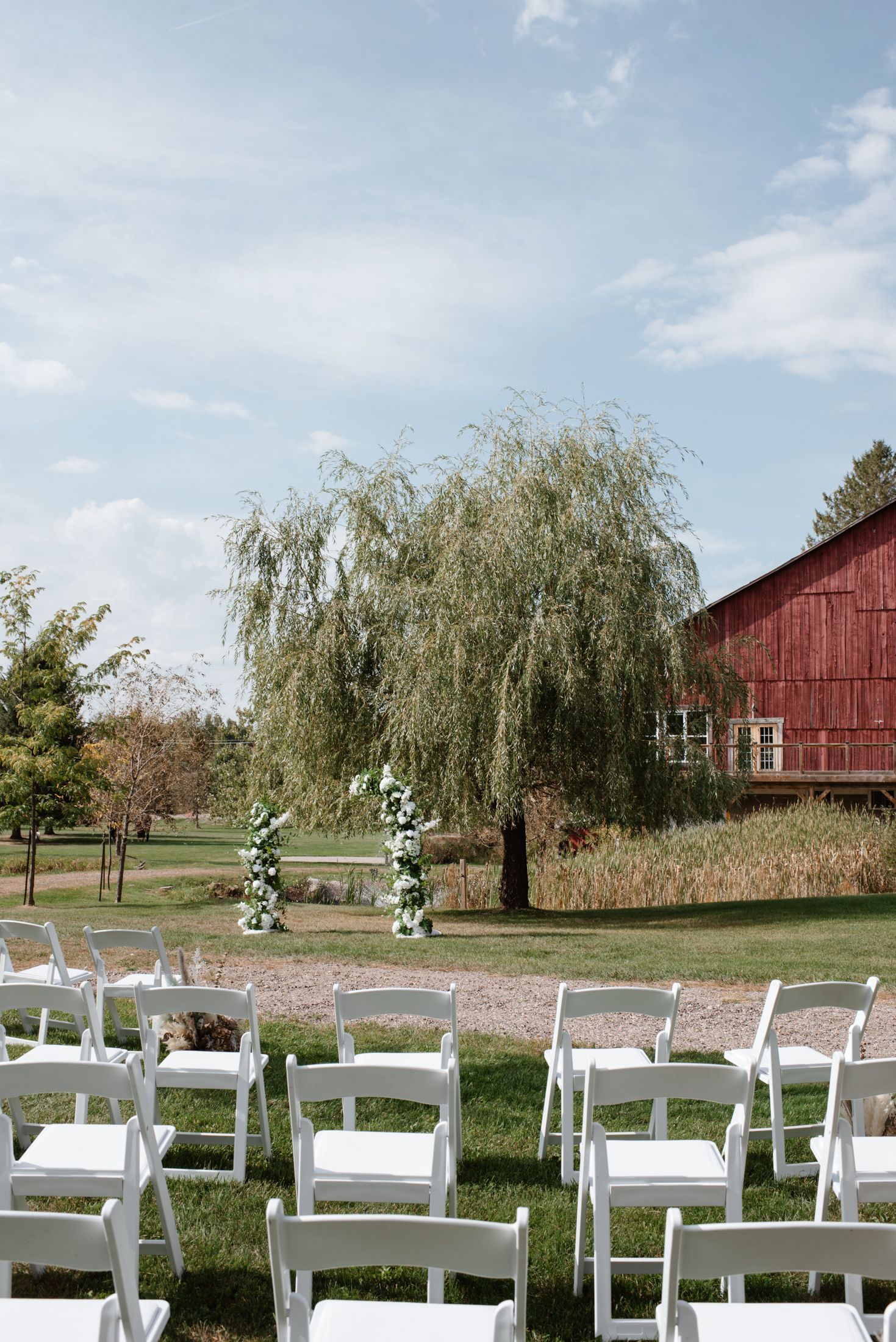 Outdoor wedding ceremony setup with white chairs, floral arch, weeping willow tree, and red barn on a sunny day.