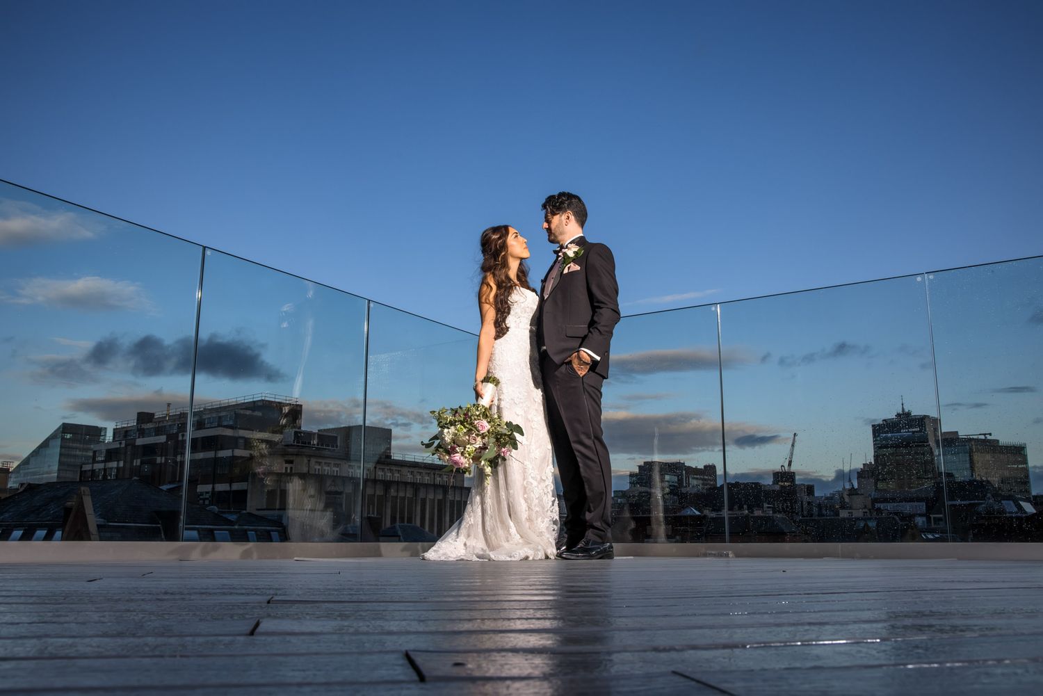 Bride and groom share a romantic moment on a rooftop terrace with glass railings and a city skyline at dusk.