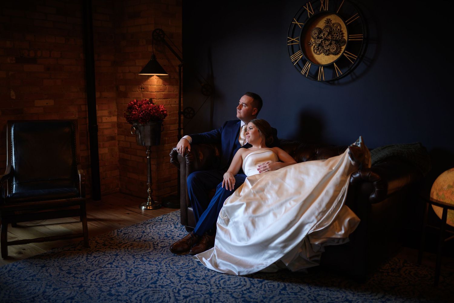 Bride and groom sitting together on a dark sofa in a moody, dimly lit room with a decorative clock on the wall.