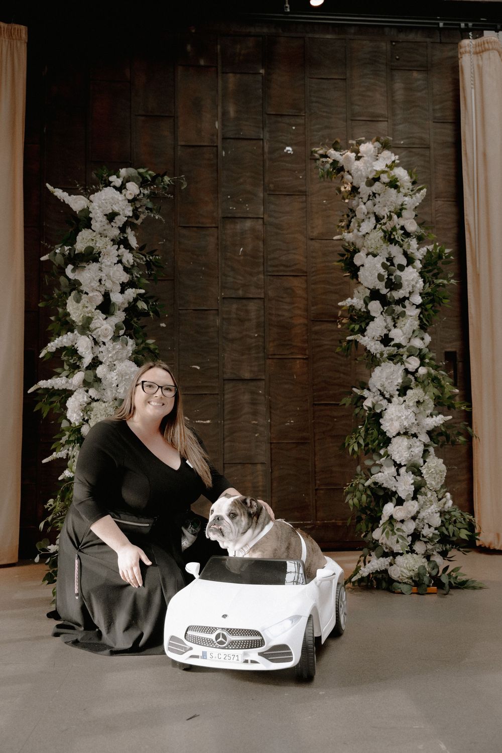 Woman in black dress poses with bulldog in toy Mercedes car under white floral arch on wooden backdrop.