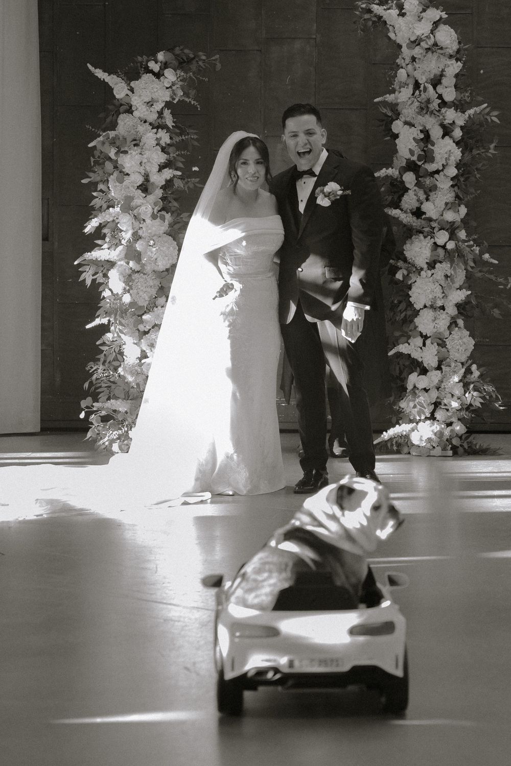 Joyful bride and groom pose at floral arch as dog rides toy car at elegant black and white wedding.