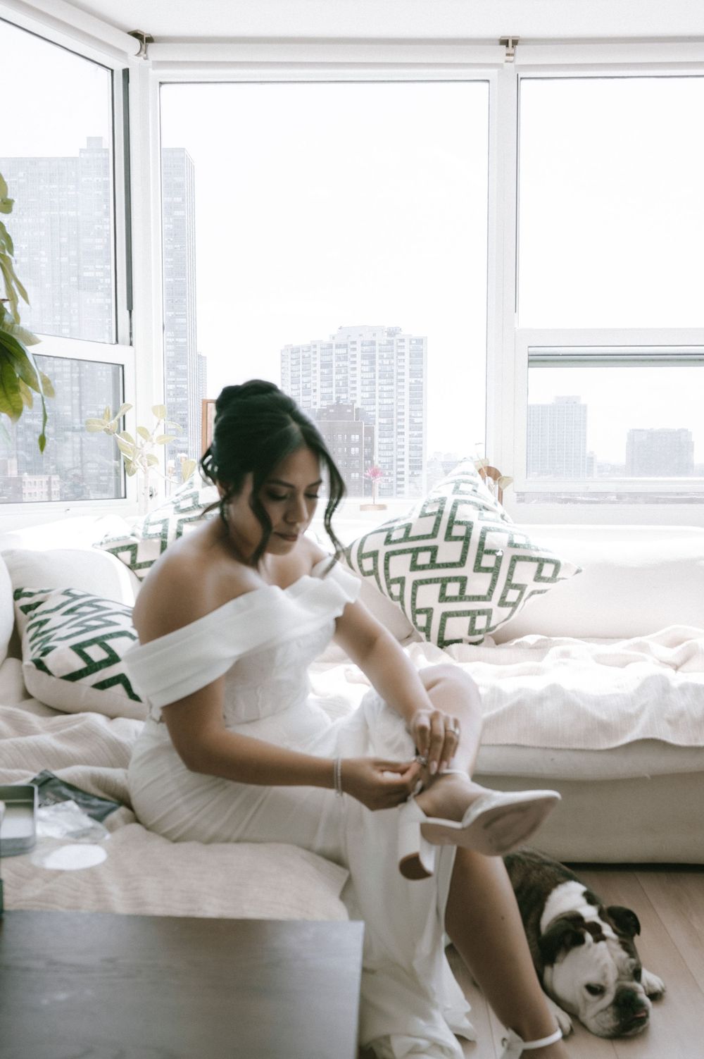 Bride in off-shoulder white gown puts on shoes while sitting on a window seat with her bulldog nearby.