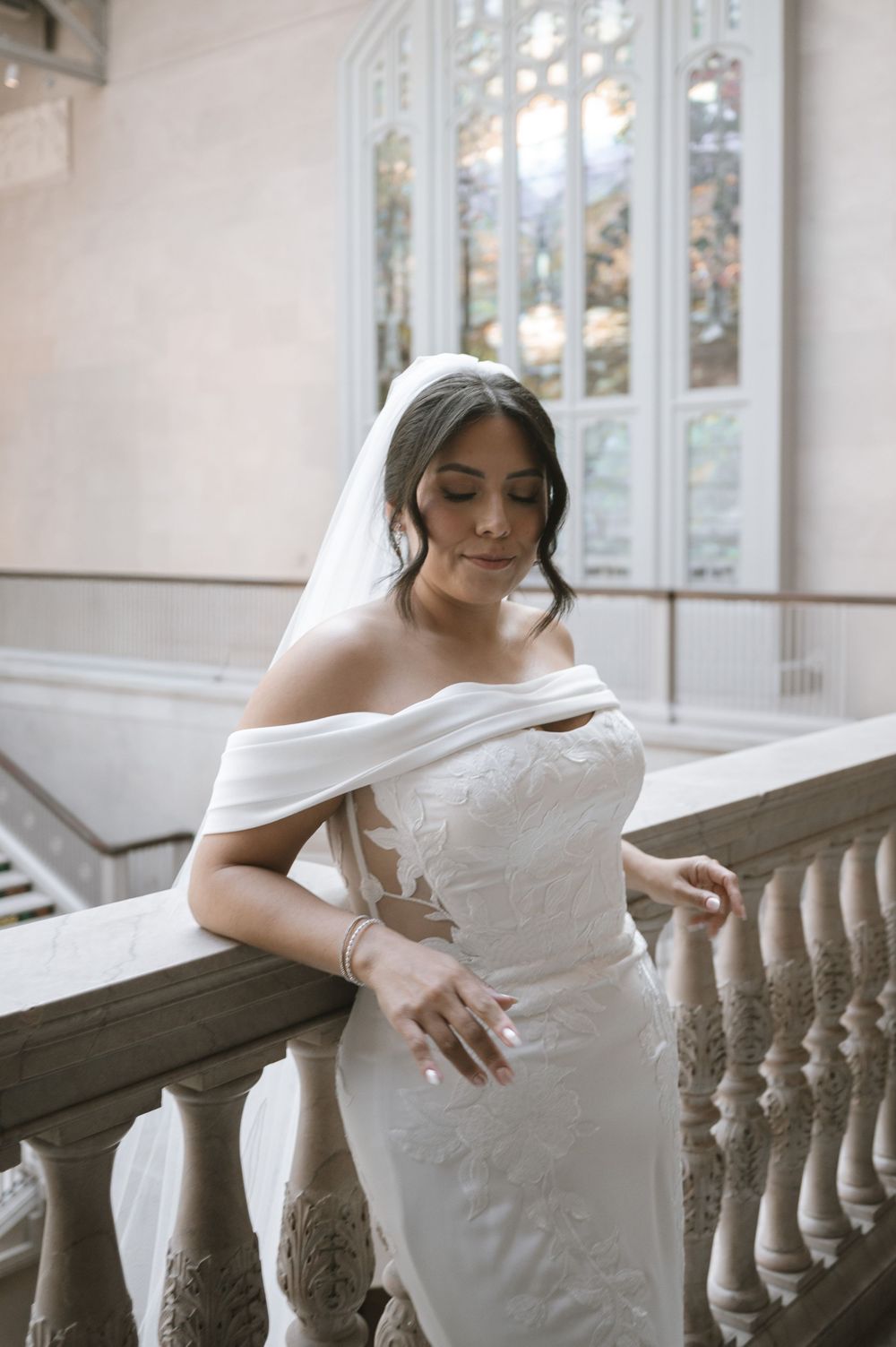 Bride in off-shoulder lace wedding gown and veil posing on ornate stone staircase.