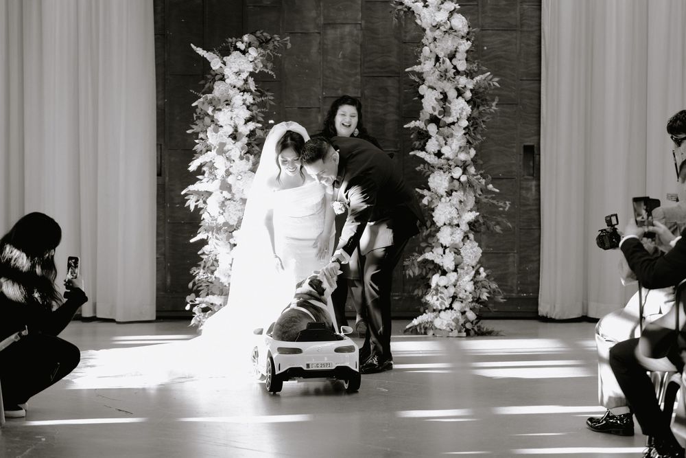 Black and white wedding photo of bride walking down aisle with dog in toy car and floral arch backdrop.