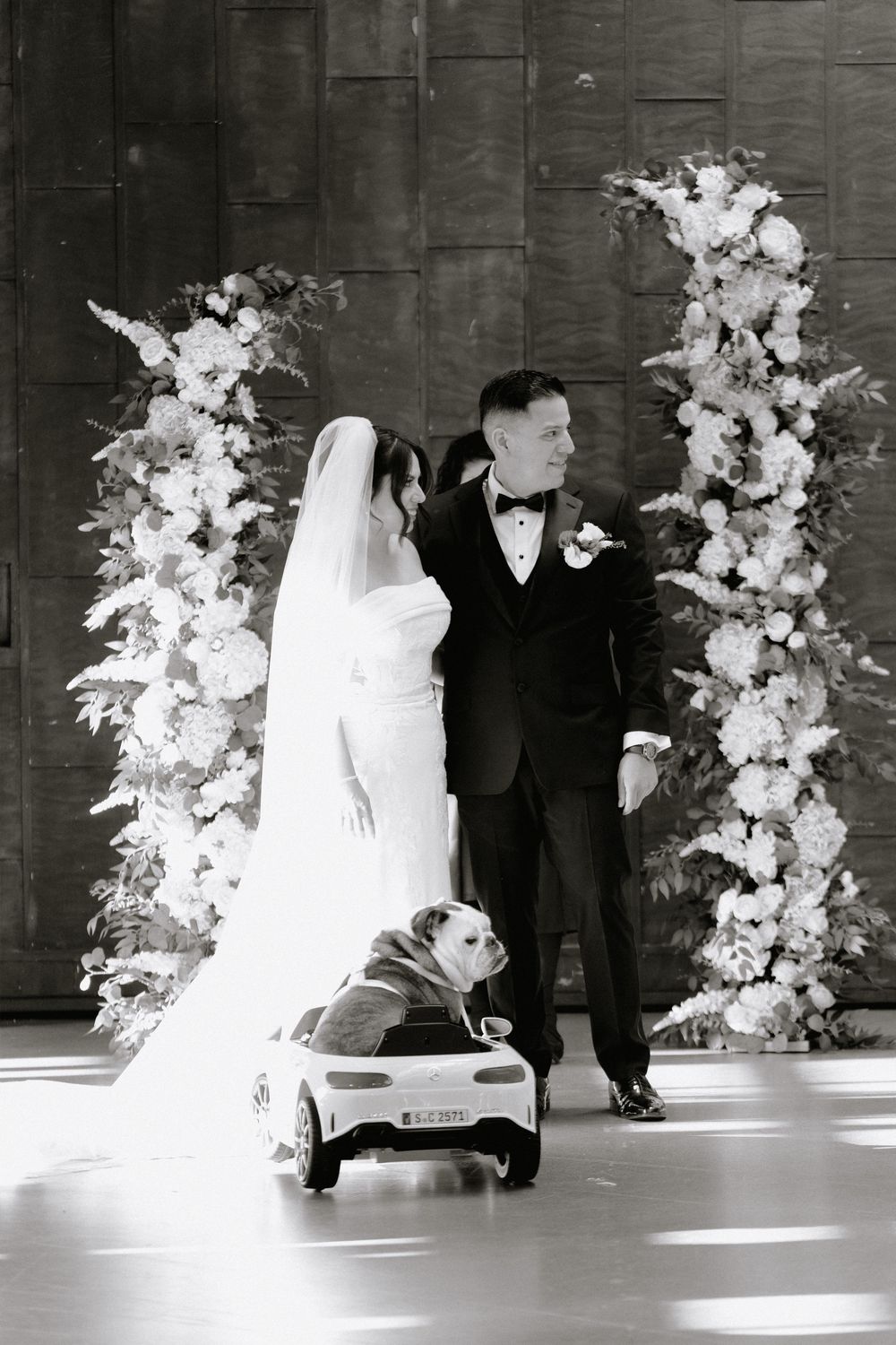 Black and white wedding photo of bride and groom with bulldog in toy car before floral arch.
