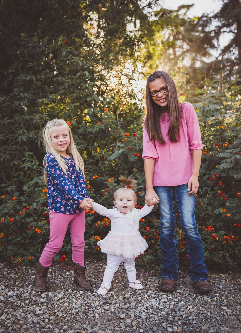 Three sisters hold hands outdoors, two older girls in pink flank a smiling baby in white dress among colorful garden foliage.