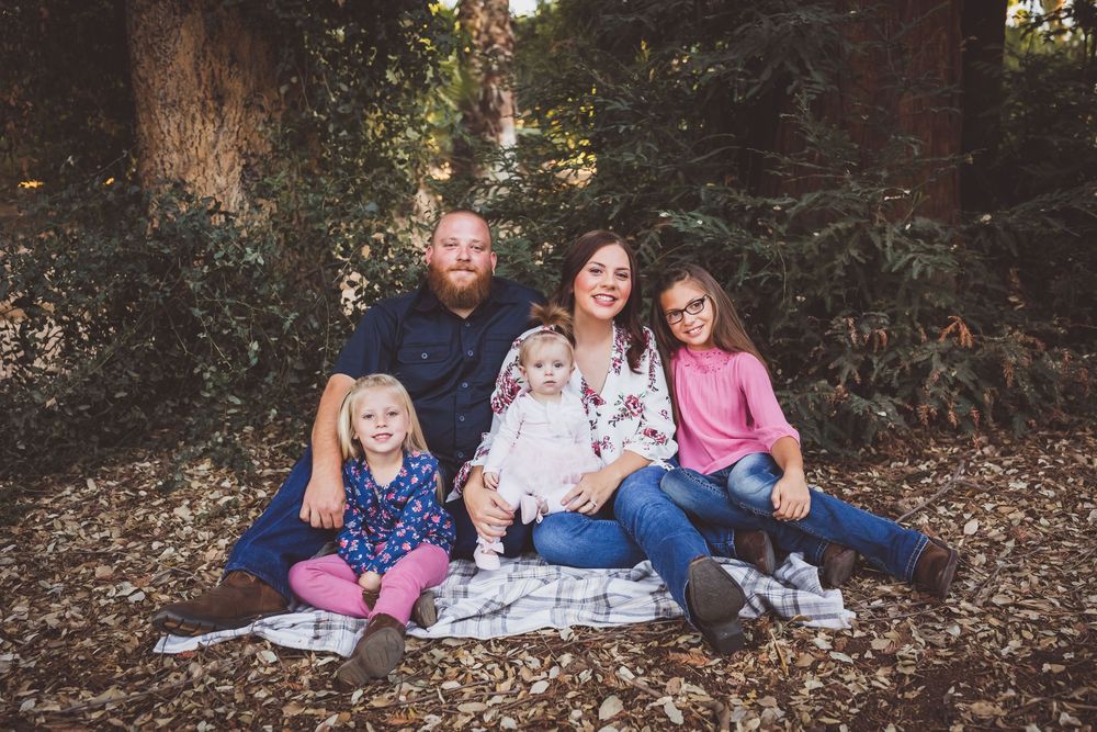 Family of five posing outdoors on autumn leaves, parents with three young daughters sitting together among trees.