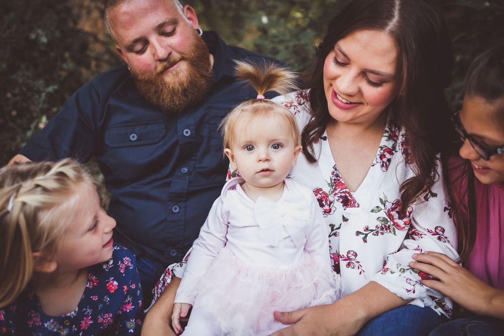 A bearded man and smiling woman sit outdoors with three children, including a baby in a pink tutu, for a family portrait.