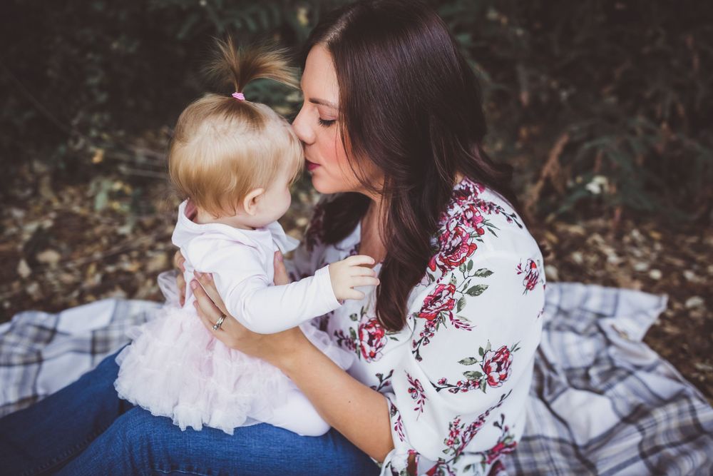 A young mother in a floral blouse holds her baby girl on a plaid blanket outdoors, sharing a tender moment.