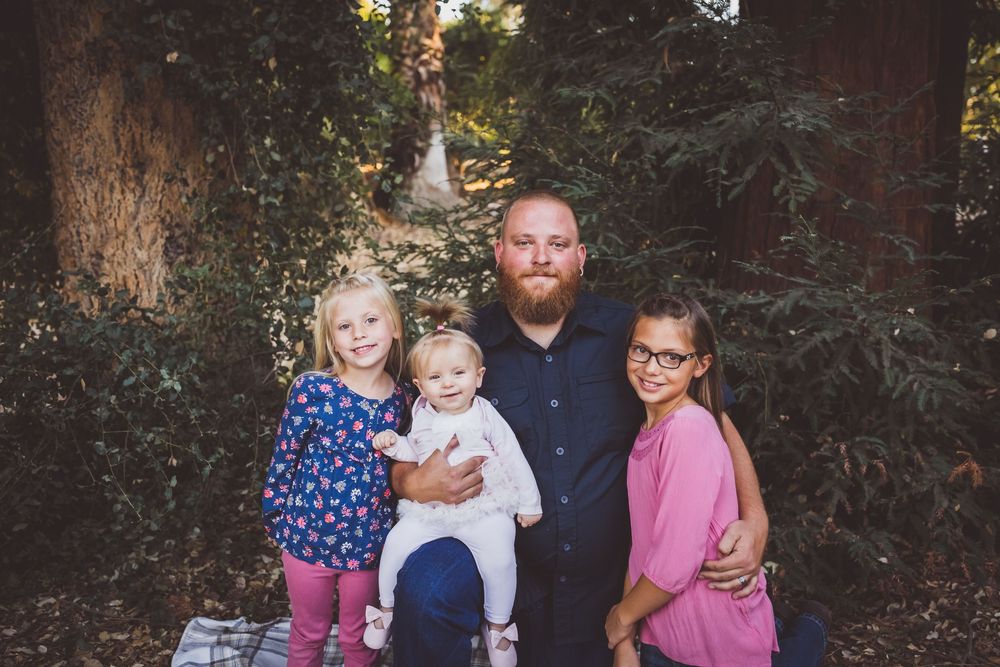 A bearded father poses outdoors with three young daughters among lush green trees in a family portrait.