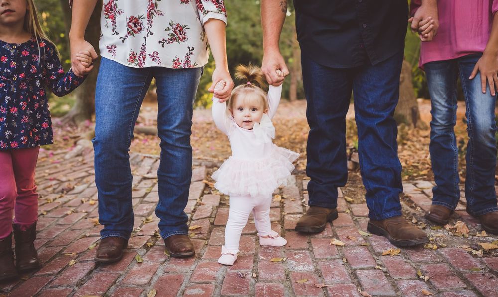 Baby girl in white dress learning to walk while family holds hands on a brick path outdoors.