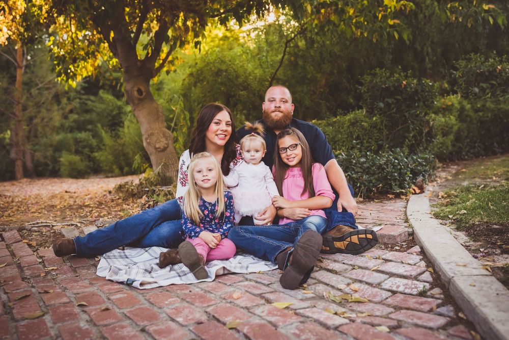 Happy family of five sitting on brick path outdoors, parents with three young daughters smiling among lush green trees.