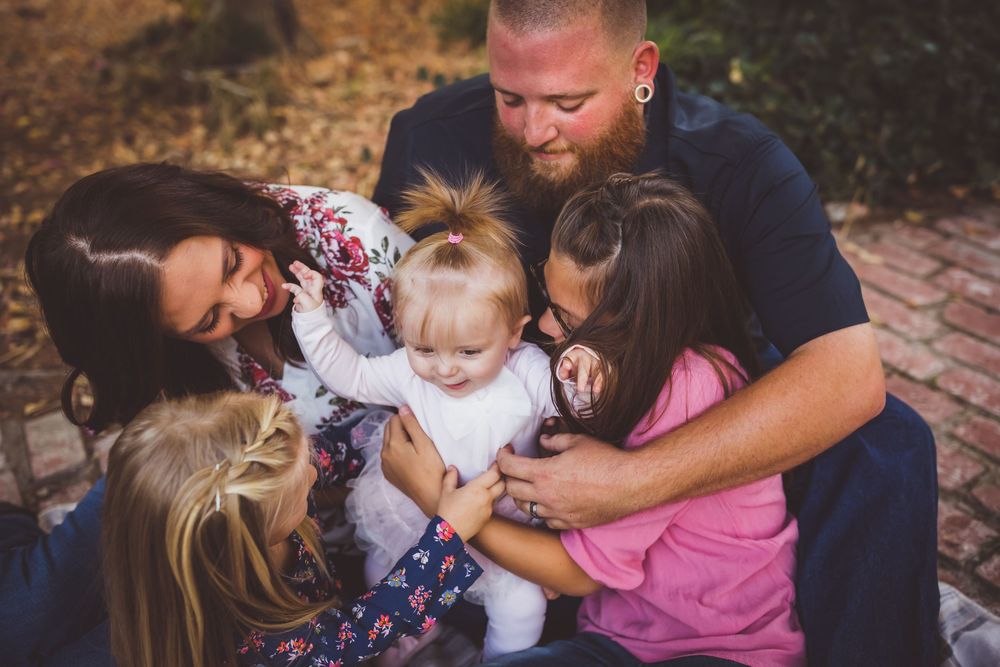 Loving family huddles together on blanket outdoors, parents and siblings embracing adorable baby girl with joy.