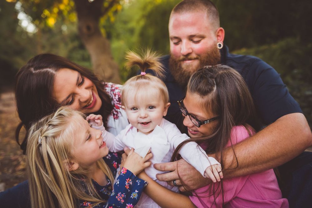 Happy family of five huddles together outdoors, parents and three daughters sharing a joyful moment with baby in white.