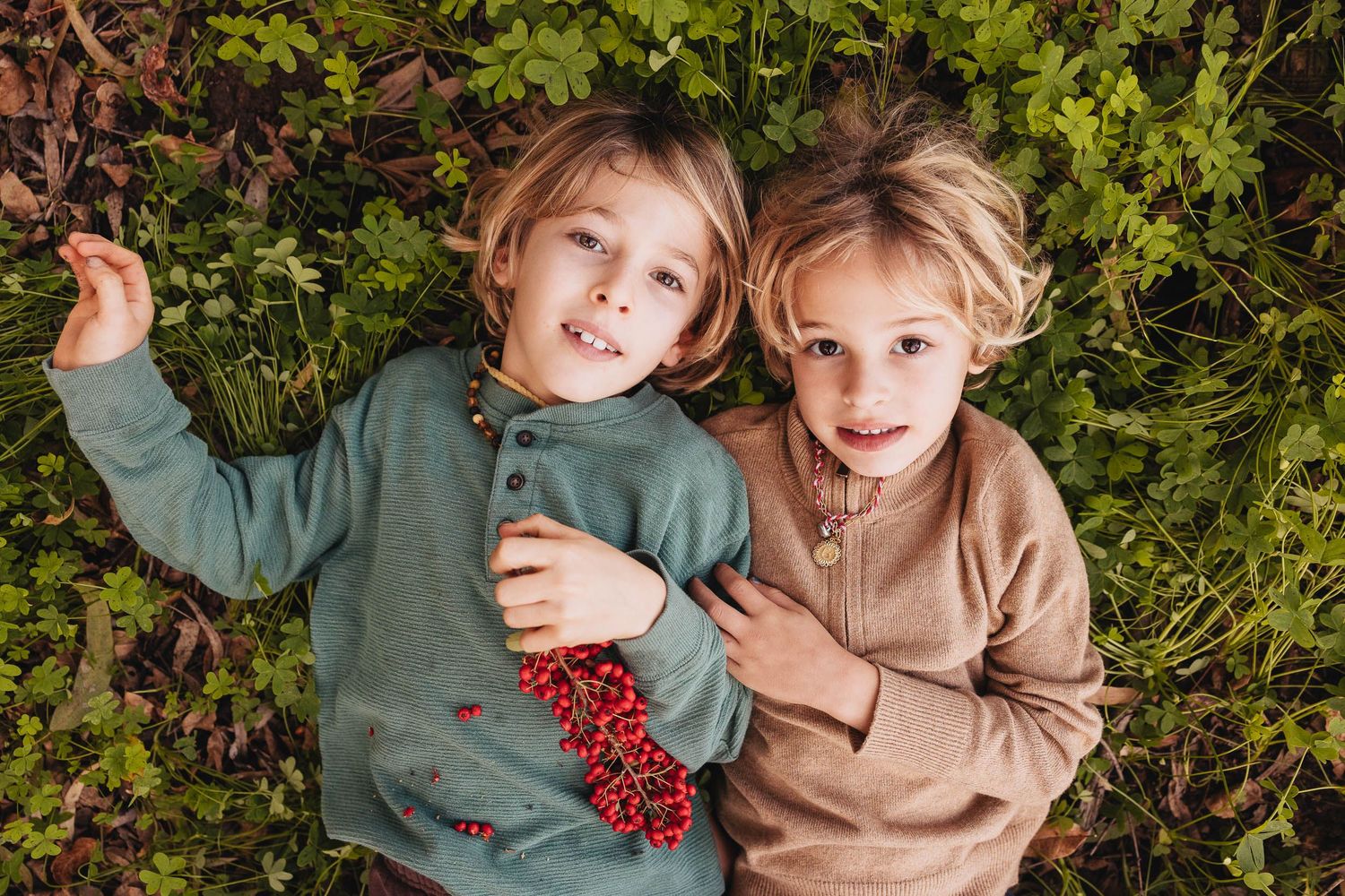 Two young children lying in lush green grass, wearing cozy sweaters, smiling up at the camera.