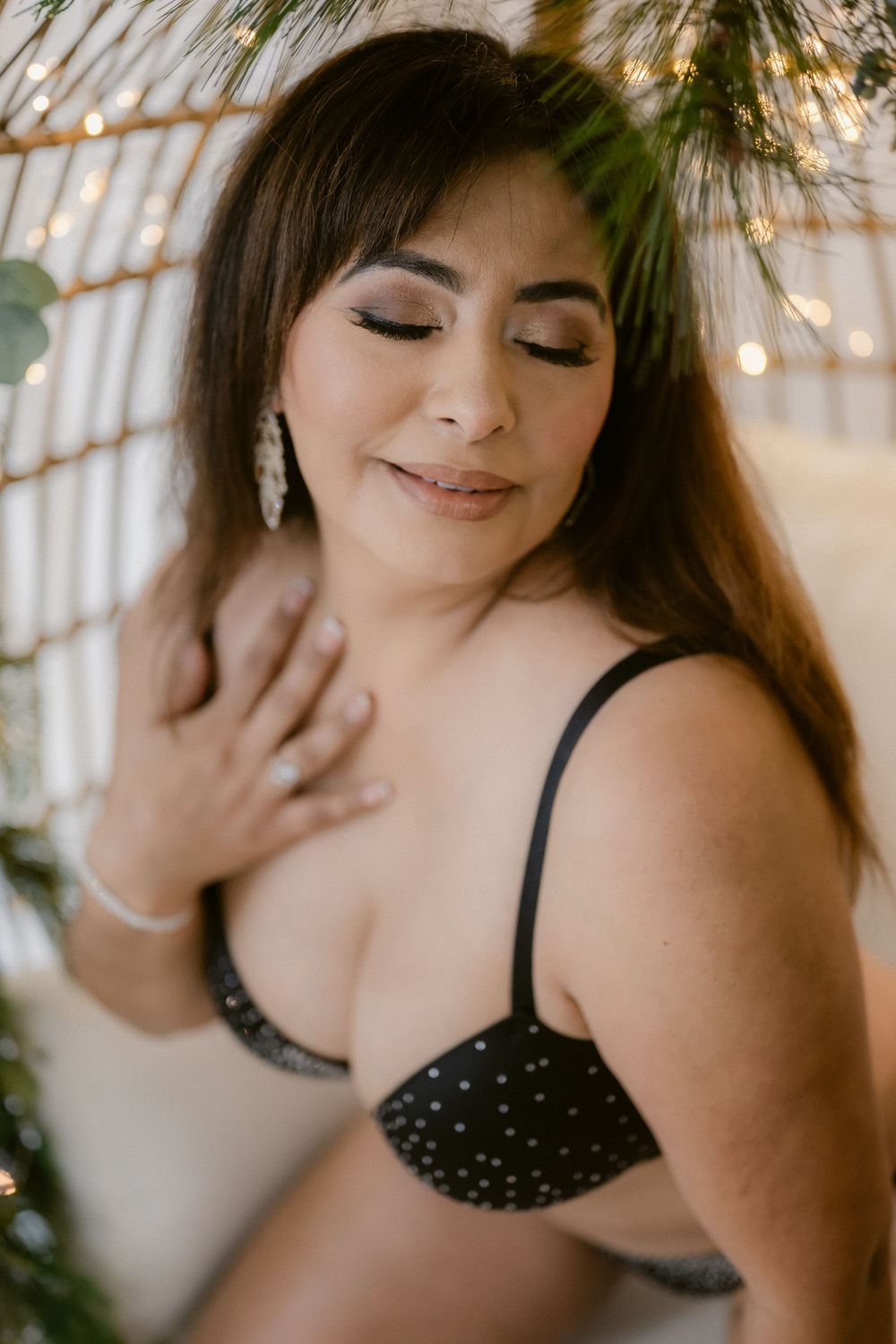 Woman in black studded bralette poses gracefully with eyes closed, surrounded by greenery and warm fairy lights.