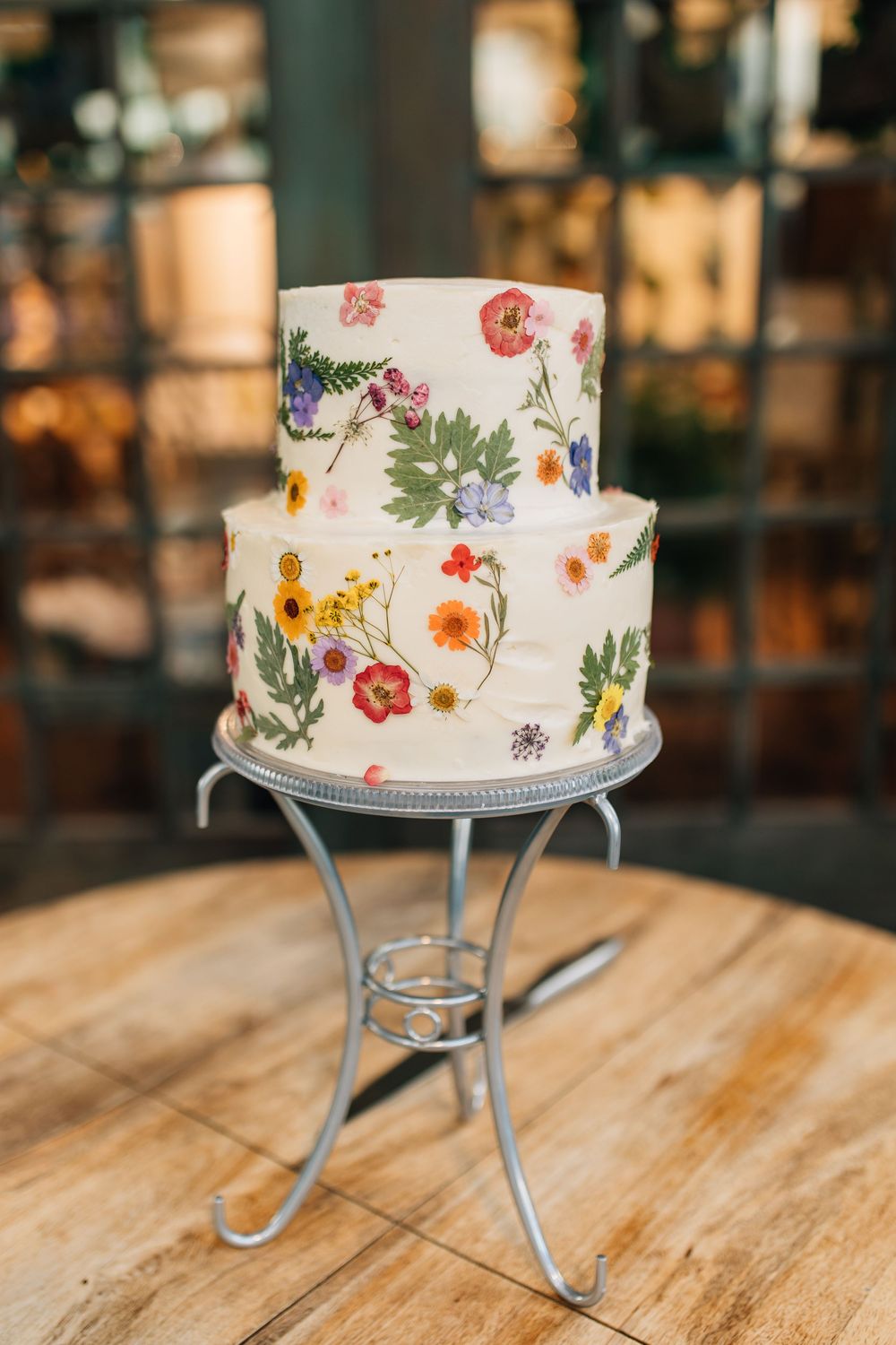 Two-tier white wedding cake decorated with pressed colorful flowers on metal stand.