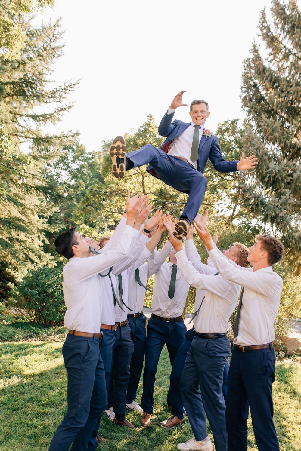 Groomsmen in white shirts tossing smiling groom in blue suit into air outdoors.