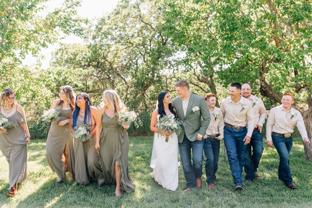Wedding party walks through green orchard, bridesmaids in olive gowns, groomsmen in khaki.