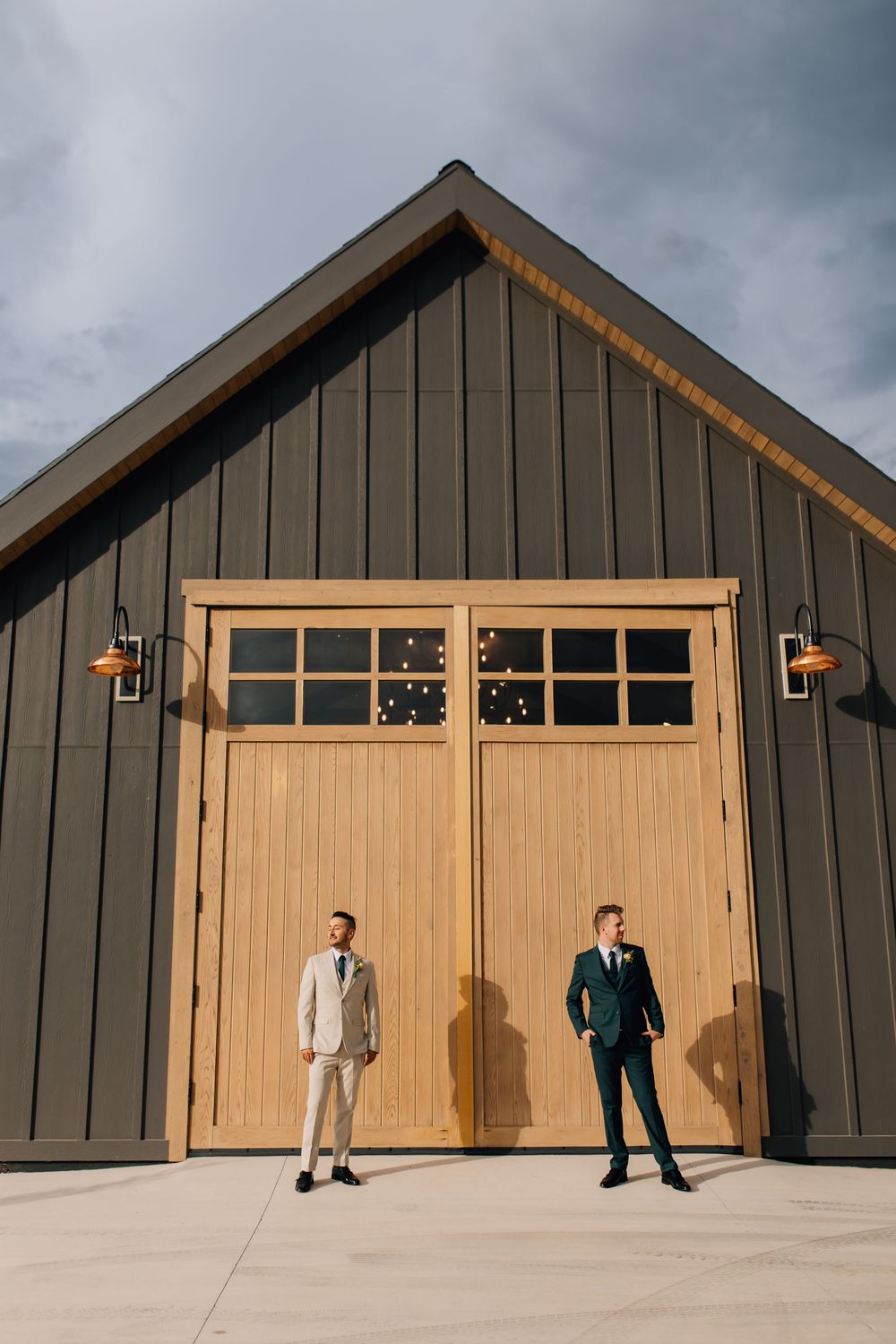 Two grooms in suits standing before a modern barn venue with large wooden doors.