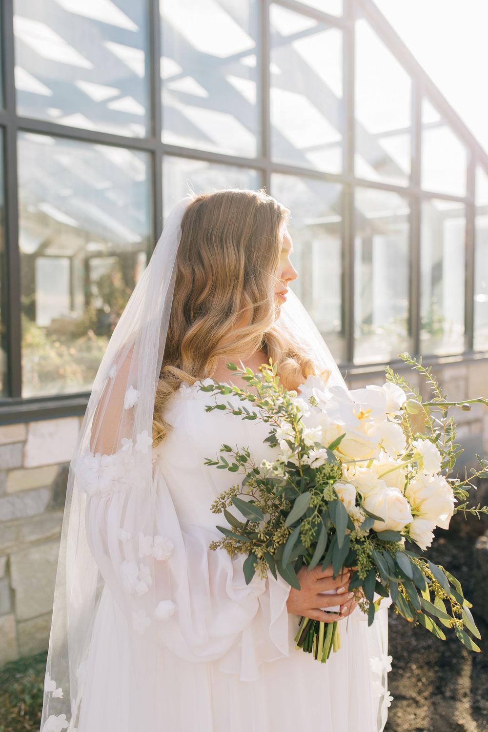 Bride in white gown holding a bouquet of white peonies and greenery in a sunlit greenhouse.