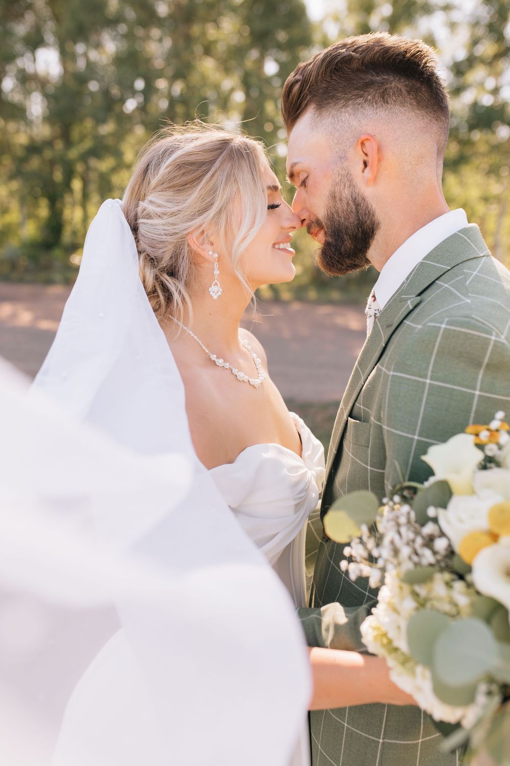 Bride and groom sharing a romantic moment outdoors, with veil flowing and greenery in the background.