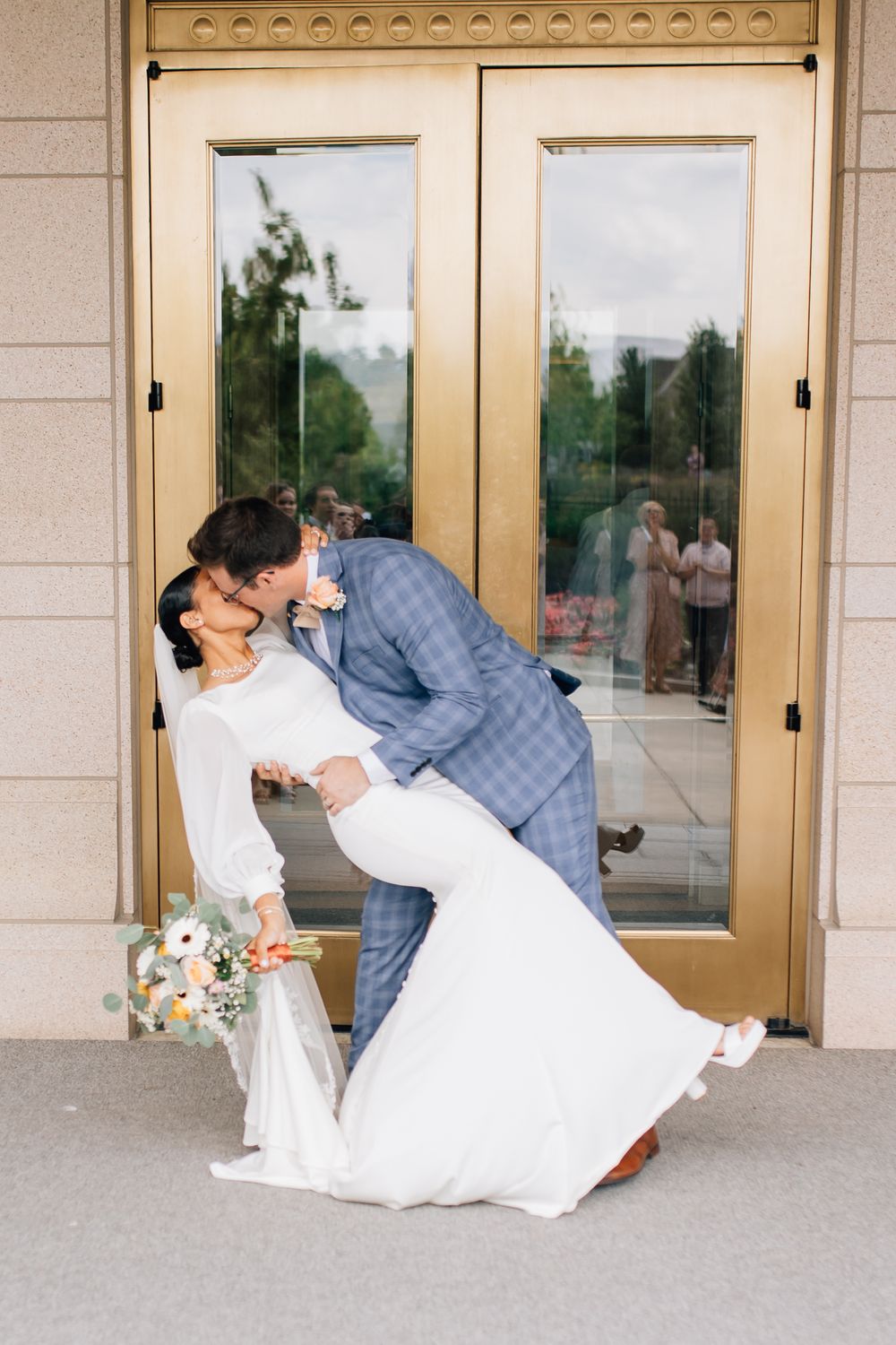 Groom dips bride for a kiss outside golden doors at elegant wedding venue.