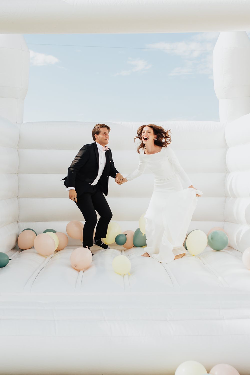 Bride and groom jump playfully in white bounce house filled with pastel balloons.