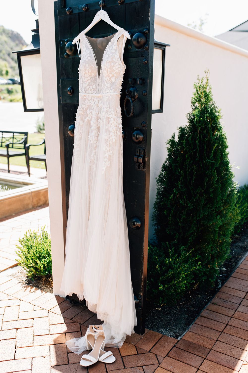 Elegant white lace wedding dress hanging on dark door with white heels below outside.