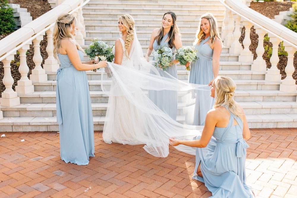 Bridesmaids in blue dresses arrange bride's veil on elegant staircase at outdoor wedding venue.