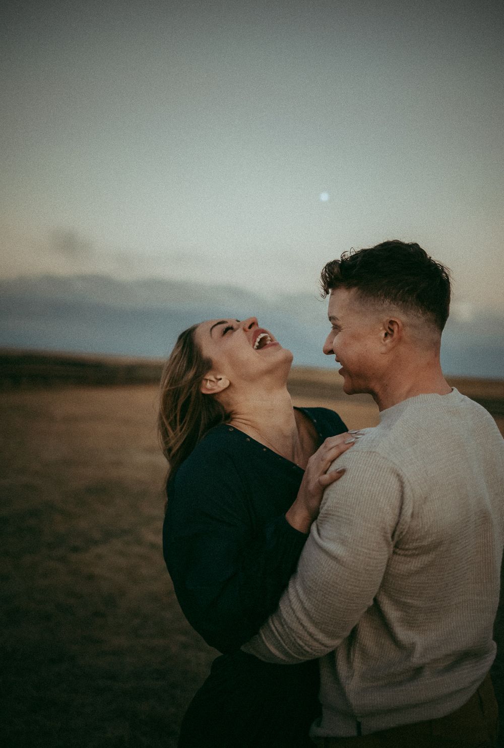 A joyful couple laughing and embracing  at dusk, with a serene twilight sky in the background.