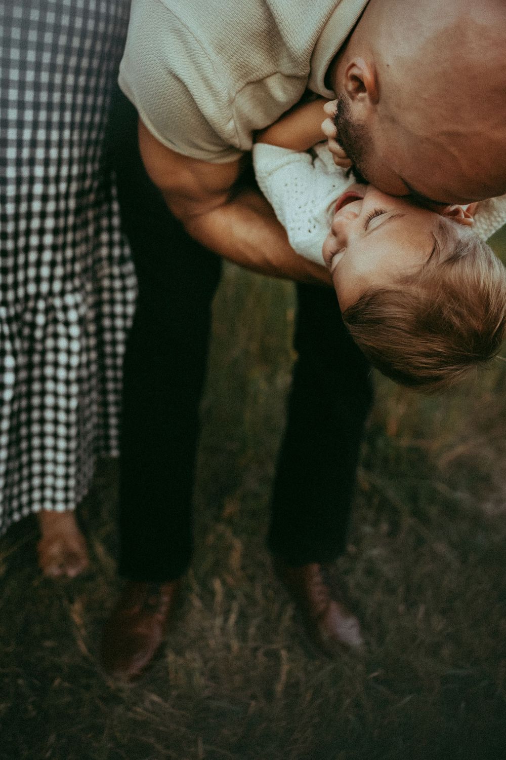 A father tenderly kisses his baby while holding them upside down outdoors in a grassy field.