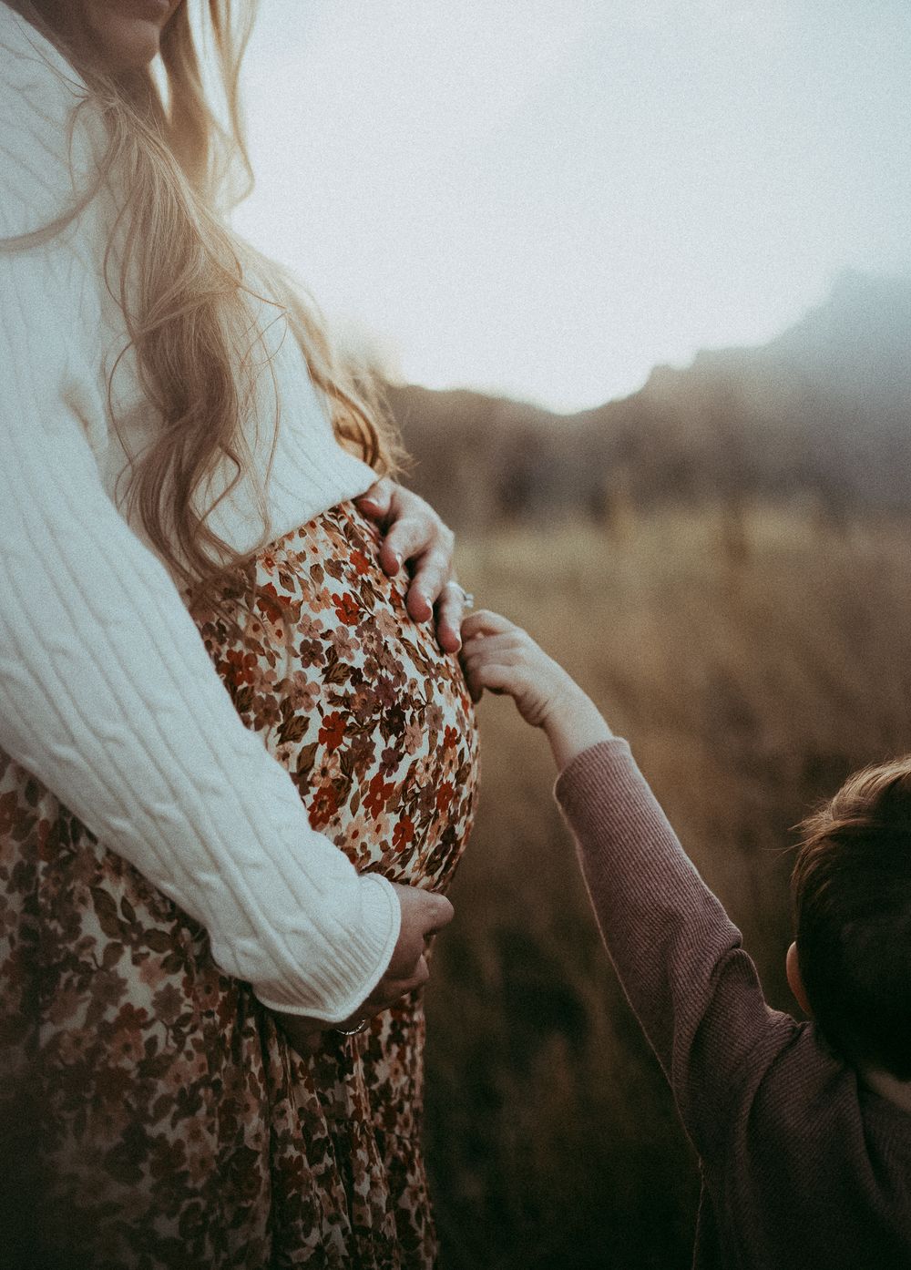 Pregnant woman in floral dress and white sweater holds child's hand in golden field at sunset.