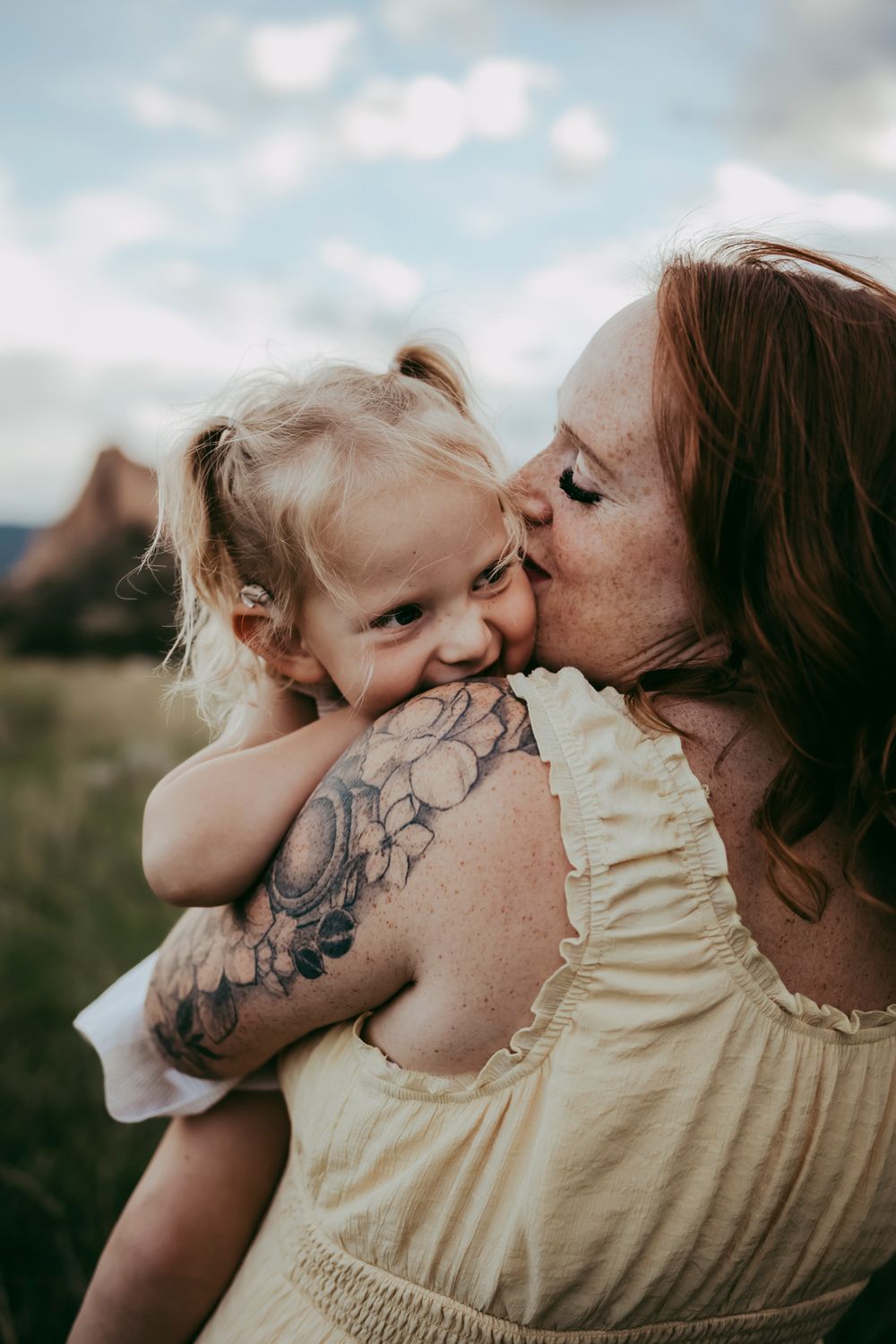 A  mother with a floral tattoo kisses her smiling toddler daughter outdoors.