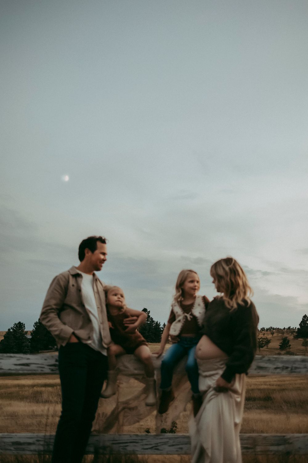 A family of four poses outdoors at dusk in a field, with a pregnant mother and father holding two young children on a fence.