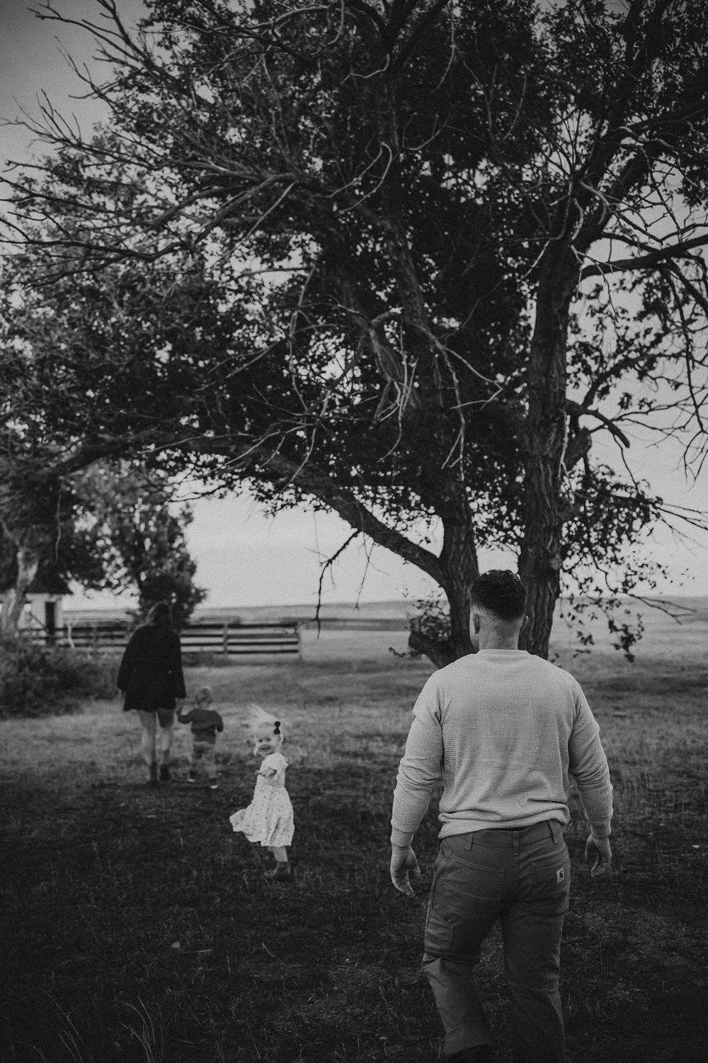 Black and white photo of a family walking toward a large tree in a rural field, with a man, woman, and small child in a dress.