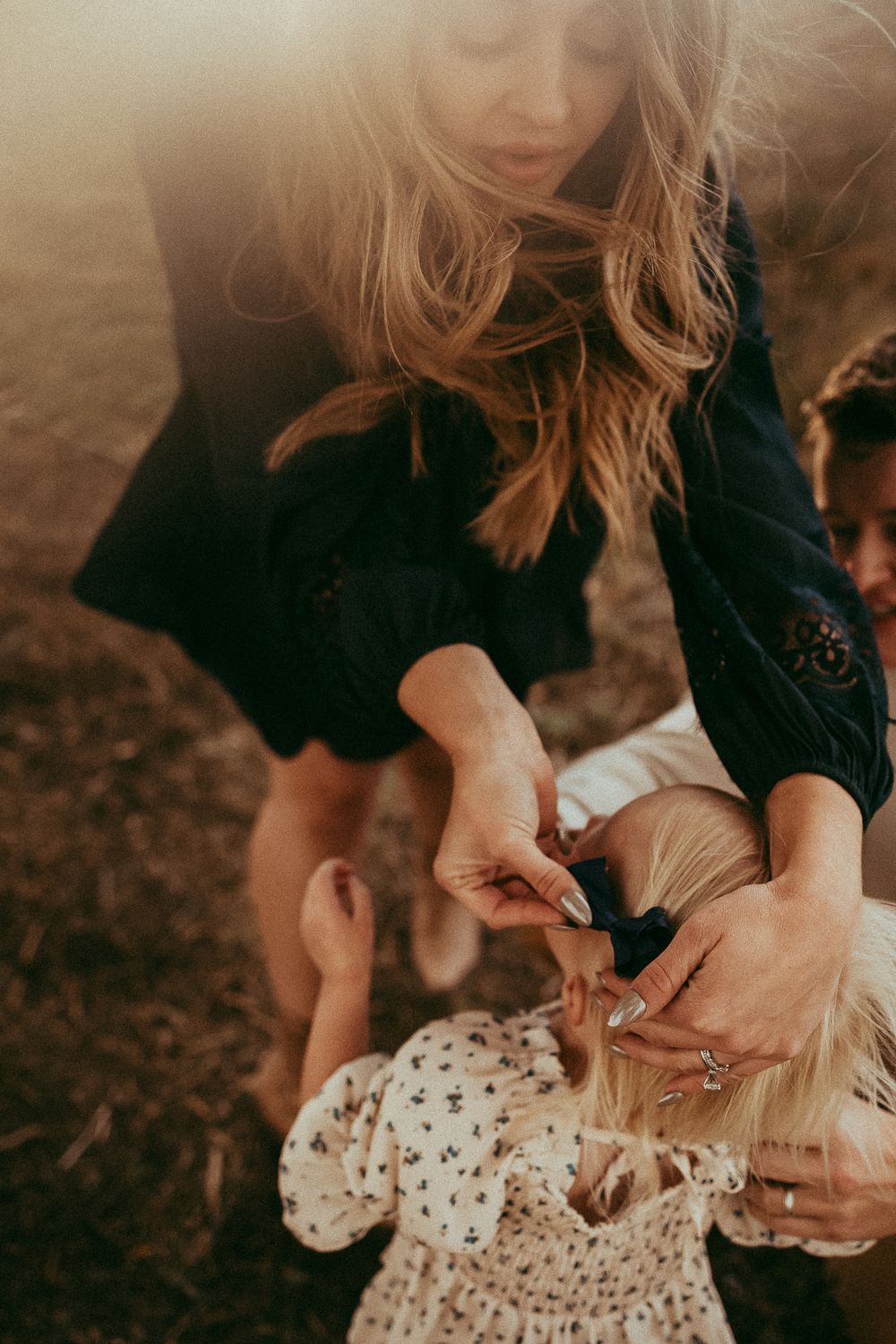 A woman with long blonde hair fixing a young girl's hair outdoors in warm golden light.