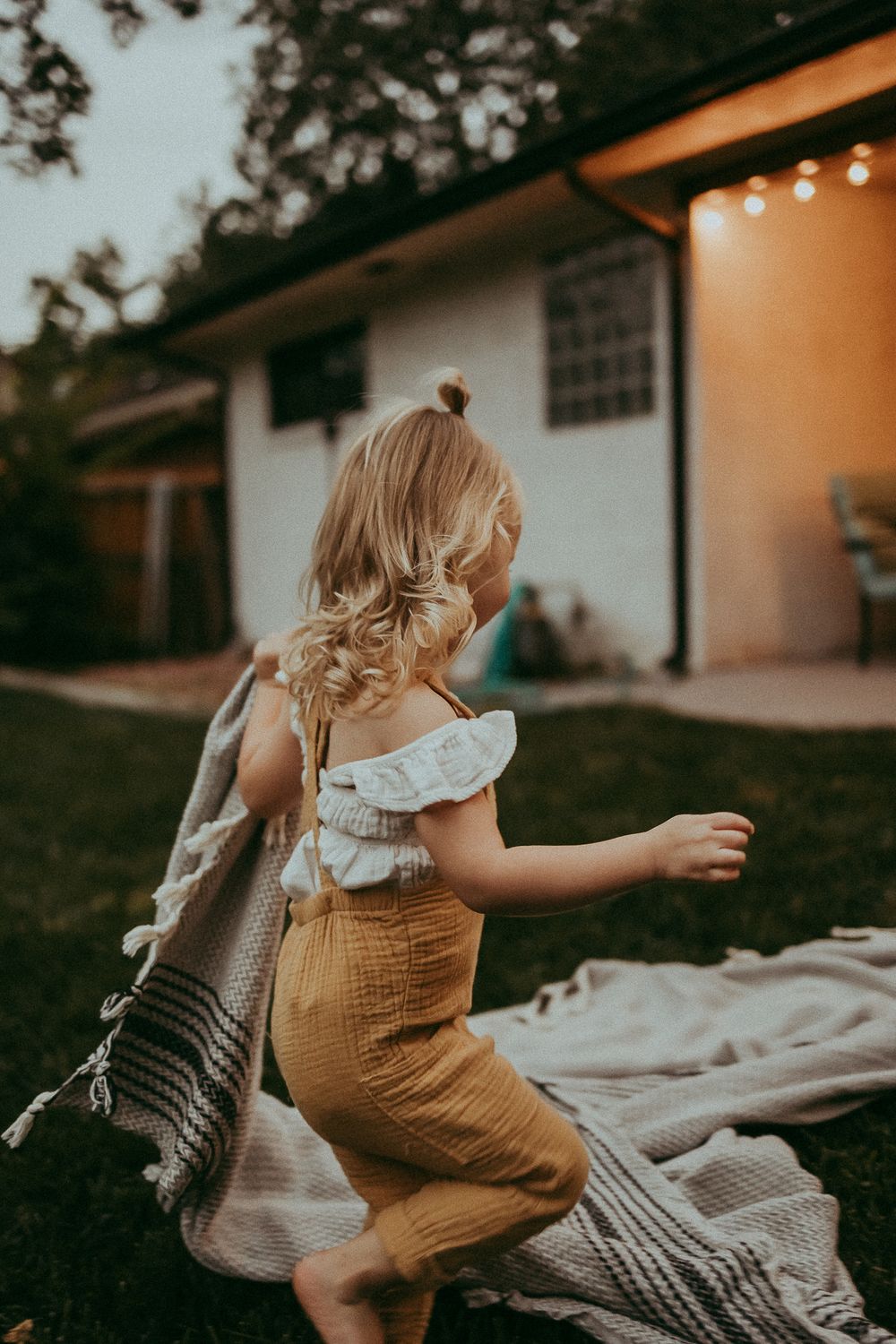 toddler girl in mustard overalls runs across a backyard lawn at dusk, dragging a striped blanket.