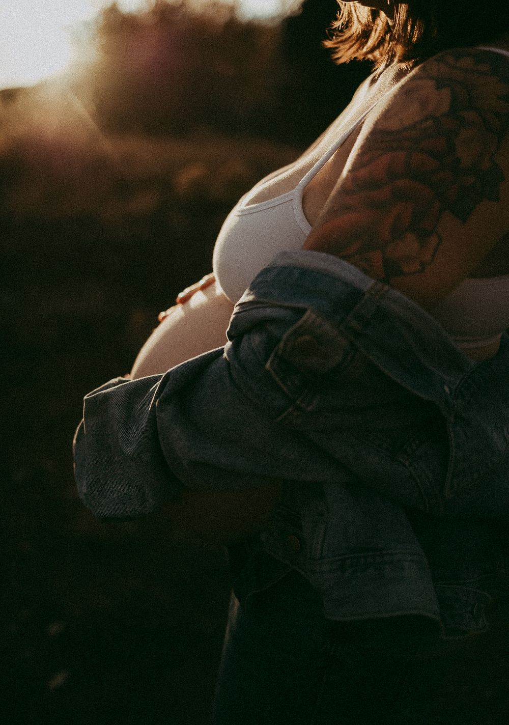 Pregnant woman with tattoos wearing a white crop top and denim jacket, glowing in golden sunset light outdoors.