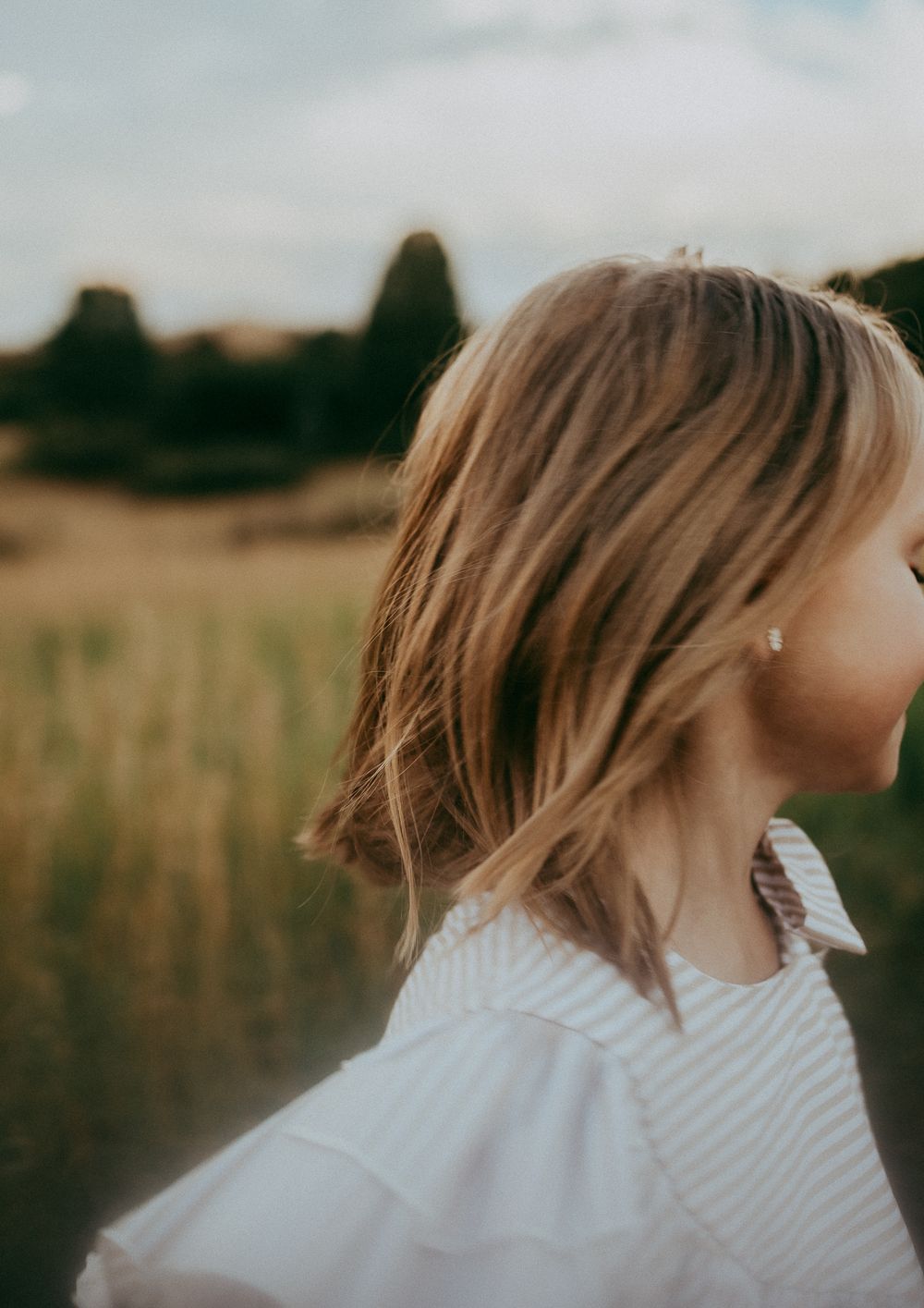 girl with blonde bob hair wearing a white ruffled top stands in a golden field at dusk.
